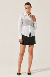 A woman is wearing a white button-up shirt with long sleeves and a collar. Over the shirt, she has layered a gray cropped knit bustier top. She is also wearing a black mini skirt and pointed black shoes.